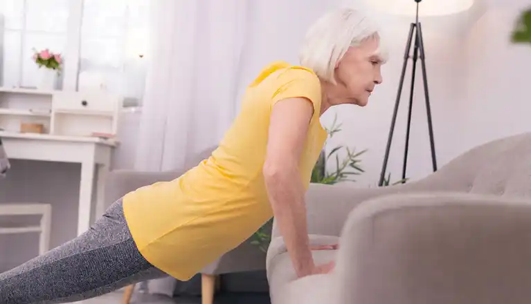 Senior woman in yellow top doing incline push-ups on a sofa at home to build strength