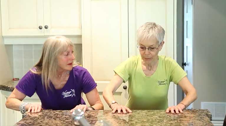 Vintage Fitness trainer Dee coaching a senior client in a home kitchen setting, demonstrating strength-building exercises for older adults
